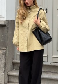 Kvinde iført en beige knapjakke og sorte bukser, bærende en sort håndtaske, stående på stentrapper foran en grå dør.