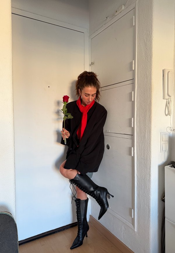 Une femme en blazer noir et en écharpe rouge tient une rose tout en ajustant une grande botte noire à lacets près de sa porte d’entrée.
