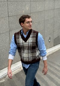Veste tricotée à carreaux marron et blanc portée sur une chemise bleu clair, avec un jean en denim, marchant sur un chemin texturé à côté d'un mur en pierre.