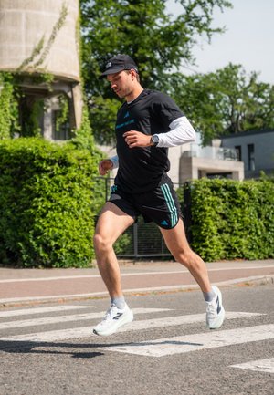 Maglietta sportiva nera con accenti blu e testo, pantaloni corti neri con strisce turchesi, scarpe da ginnastica bianche. Correndo su un attraversamento pedonale con sfondo di vegetazione.