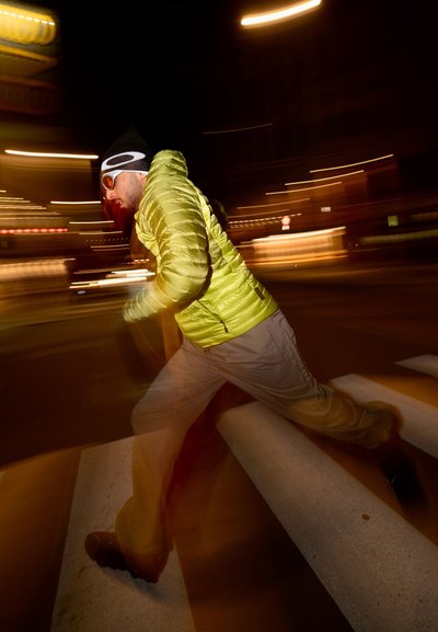 Chaqueta de pluma verde neón, pantalones gris claro y zapatos negros. Persona cruzando un paso de peatones, con luces de la ciudad desenfocadas al fondo.