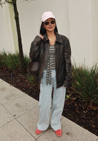 Mujer con chaqueta de cuero negra, camiseta a rayas, vaqueros azul claro, gorra rosa, gafas de sol redondas y zapatos rojos, de pie en una acera cerca de plantas.