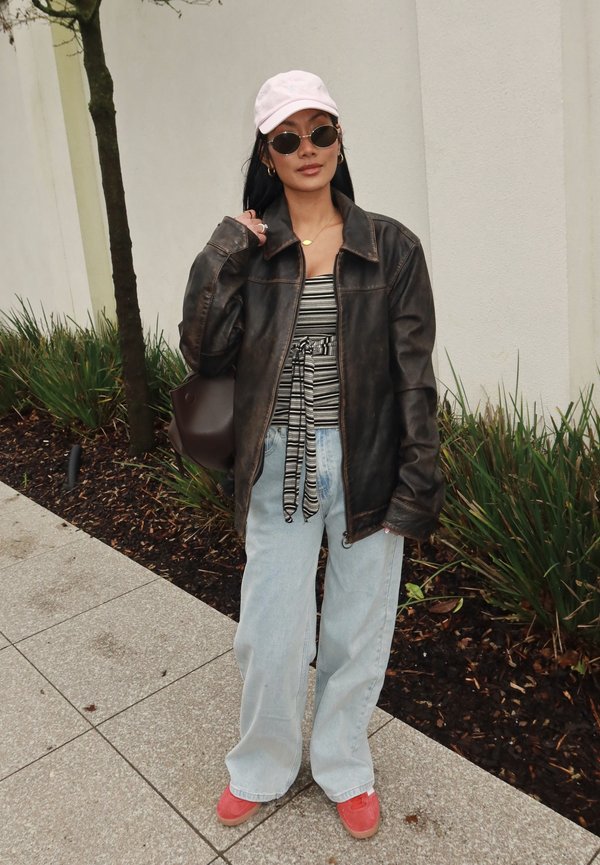 Kvinde iført sort læderjakke, stribet top, lyseblå jeans, lyserød kasket, runde solbriller og røde sko, stående på en fortov nær planter.