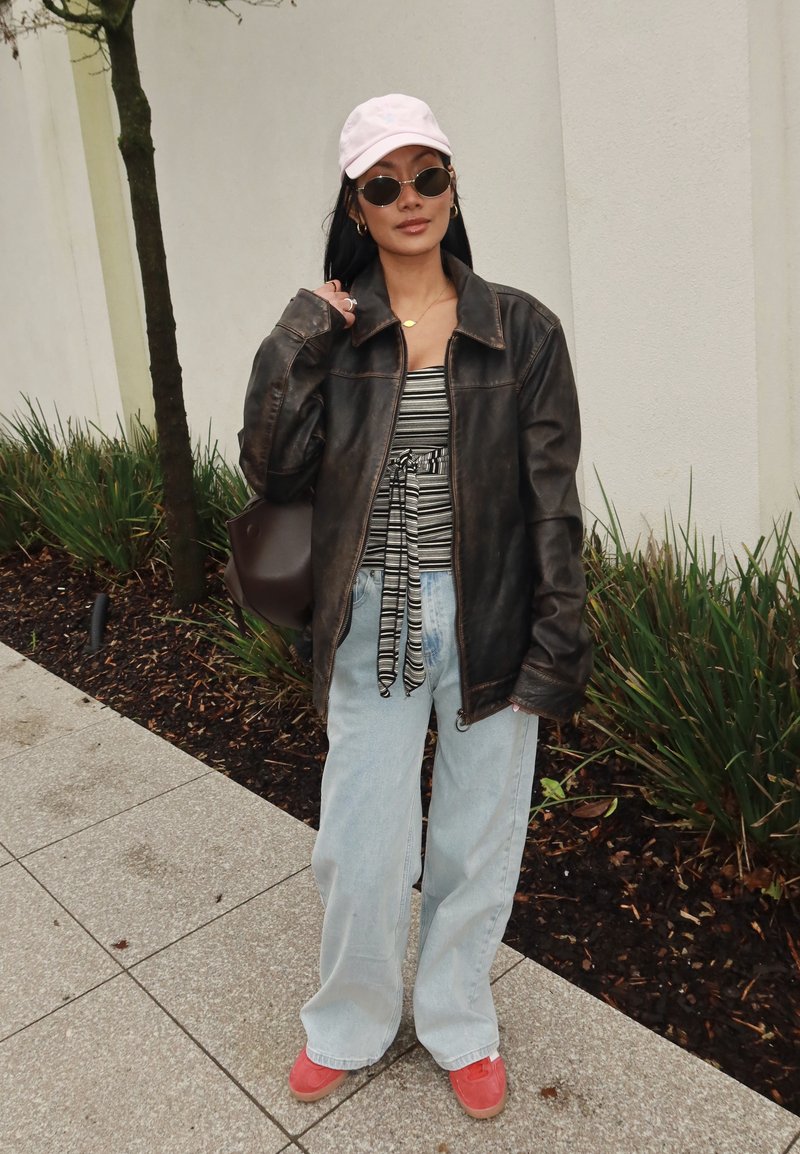 Žena v černé kožené bundě, pruhovaném topu, světle modrých džínách, růžové čepici, kulatých slunečních brýlích a červených botách stojí na chodníku u rostlin.