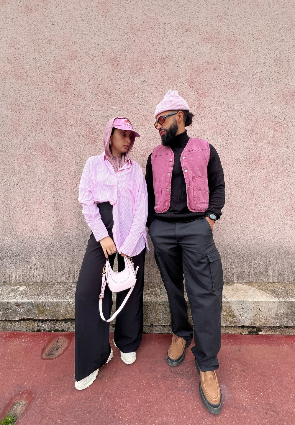 Ζευγάρι που στέκεται δίπλα-δίπλα. Ο ένας φοράει ένα ανοιχτό ροζ ριγέ πουκάμισο και φαρδιά μαύρα παντελόνια, κρατώντας μια μίνι ροζ τσάντα. Ο άλλος φοράει ένα σκούρο γιλέκο πάνω από μαύρο φανελάκι και cargo παντελόνια.