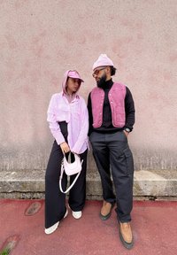 Ζευγάρι που στέκεται δίπλα-δίπλα. Ο ένας φοράει ένα ανοιχτό ροζ ριγέ πουκάμισο και φαρδιά μαύρα παντελόνια, κρατώντας μια μίνι ροζ τσάντα. Ο άλλος φοράει ένα σκούρο γιλέκο πάνω από μαύρο φανελάκι και cargo παντελόνια.