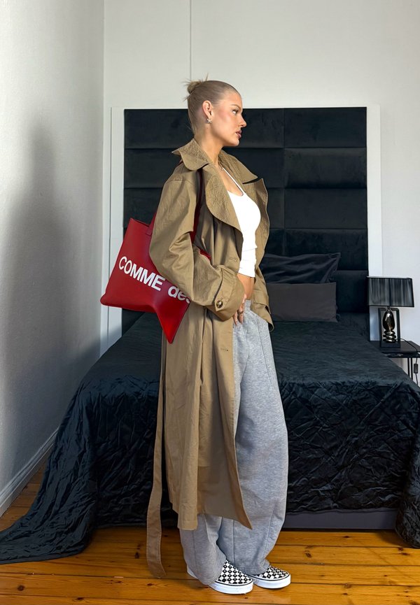 Donna con trench beige, top corto bianco, pantaloni della tuta grigi, scarpe slip-on a scacchi, che porta una grande borsa rossa, in piedi nella camera da letto accanto al letto.