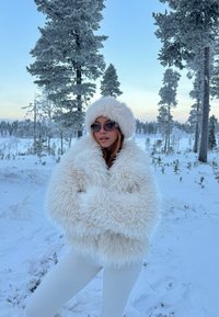 Luddig vit pälsrock och matchande hatt, båda med mjuk textur. Modellen bär mörka solglasögon mot en bakgrund av en snöig skog.