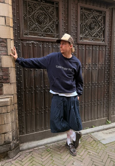 Hombre con sudadera azul marino, pantalones cortos sueltos y gorra apoyado en una pared de ladrillos, de pie en una calle adoquinada frente a una gran puerta de madera ornamentada.
