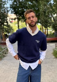Jūras spēku adīts džemperis ar V veida kakla izmēru un krēmkrāsas apkakli virs baltām pogām. Apvienojiet ar ziliem džinsiem; sudraba pulkstenis redzams uz plaukstas.