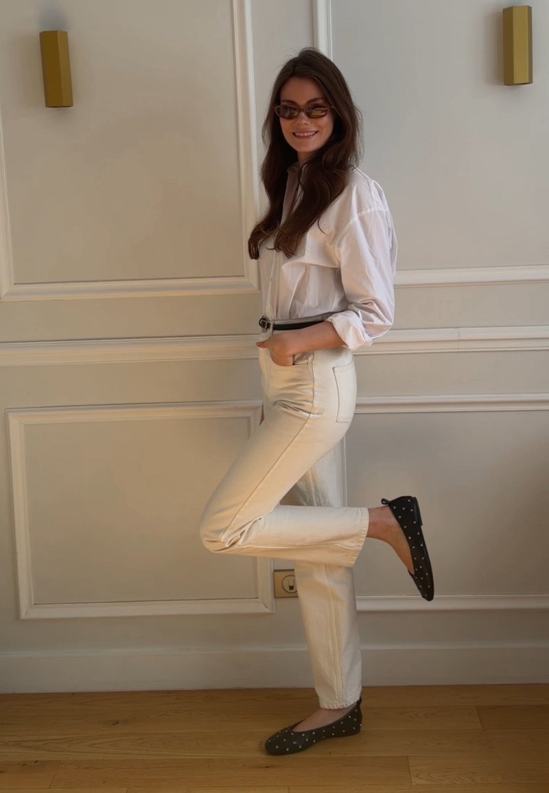 Femme portant des lunettes de soleil, une chemise blanche, un pantalon crème et des ballerines noires à pois, debout à l'intérieur avec une jambe pliée derrière elle.