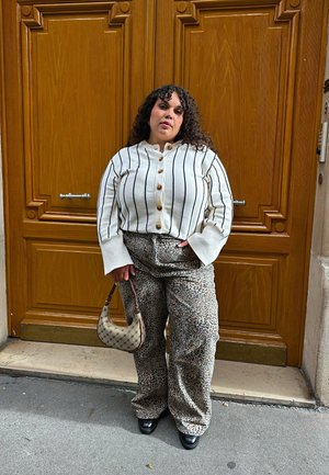 Cardigan blanco a rayas con botones y pantalones de leopardo beige. Zapatos de tacón negros y un bolso beige con asa. Fondo de puerta de madera.