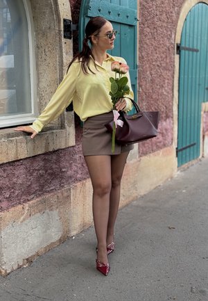 Camisa amarilla de manga larga, mini falda marrón, tacones rojos de charol con detalles, sosteniendo rosas rosas, de pie junto a una pared de piedra texturizada y puertas azules.