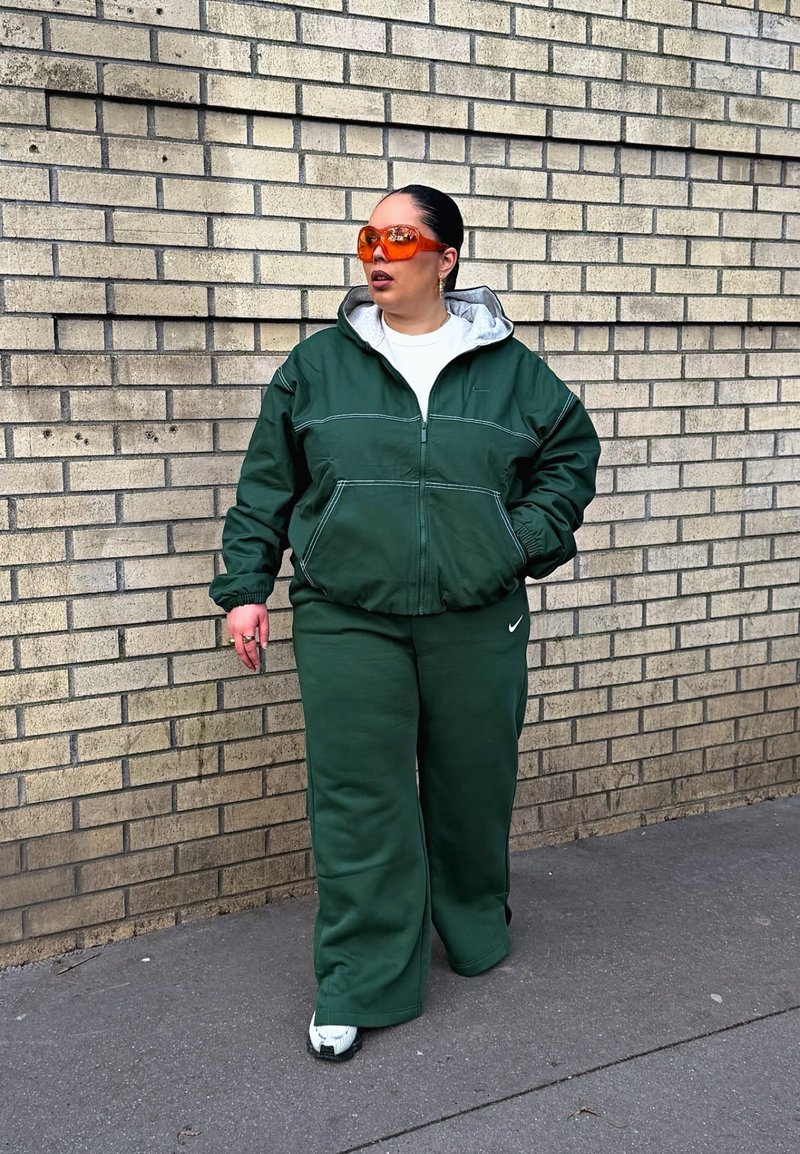 Personne portant un survêtement Nike vert foncé et de grandes lunettes de soleil orange marchant sur le trottoir à côté d'un mur en briques claires.