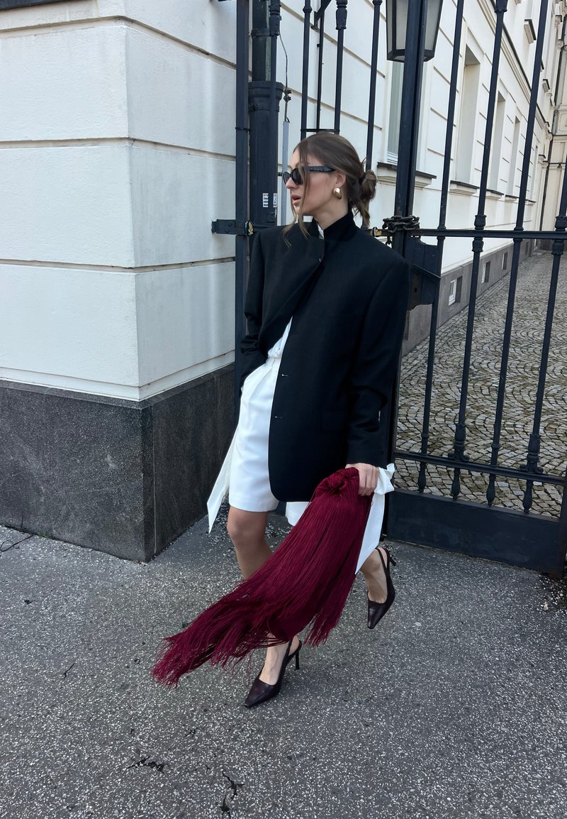 Frau trägt einen übergroßen schwarzen Blazer über einem weißen Kleid, schwarze Absätze, Sonnenbrille, goldene Ohrringe, hält eine lange rote Fransen-Clutch und steht neben einem Eisentor.