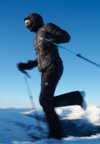 Person i sort vinterudstyr og goggles, der står på ski med stave på en snedækket bjerg under klar blå himmel.