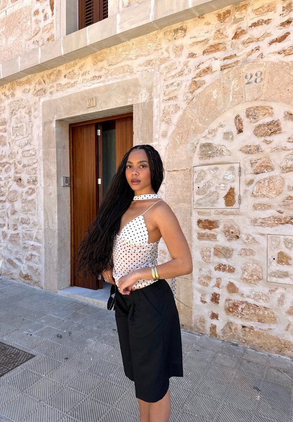 Femme aux longs cheveux tressés portant un haut blanc à pois et un short noir, debout dans une rue pavée près d'un bâtiment en pierre.