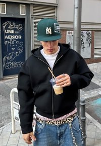 Mustapaneedega kapuutsiga särk, valge t-särk, sinised teksad, leopardimustriga vöö, roheline müts logoga. Hoides jäälattega kohvi läbipaistvas topsis.