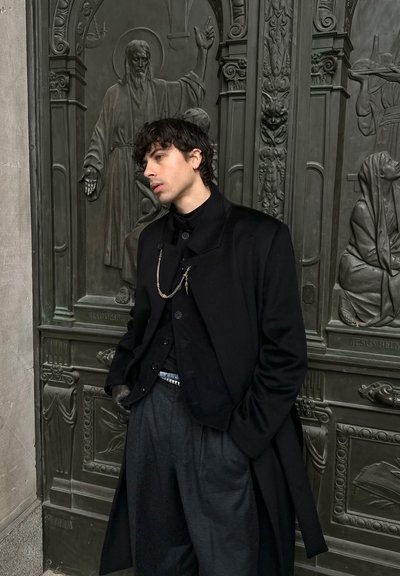 Joven con abrigo negro y guantes apoyado contra una puerta de bronce ornamentada con relieves escultóricos religiosos, con las manos en los bolsillos, mirando de lado.