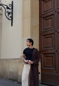 Man med tatueringar i svart skjorta och vita byxor håller solglasögon och kaffe stående vid en stor träport och beige stenvägg.