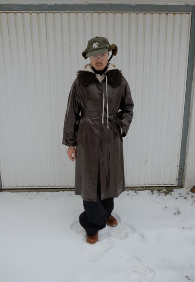 Persona con abrigo marrón oscuro con cinturón y cuello de piel, gorro de invierno verde con orejeras, gafas, de pie sobre la nieve frente a una puerta de garaje blanca.