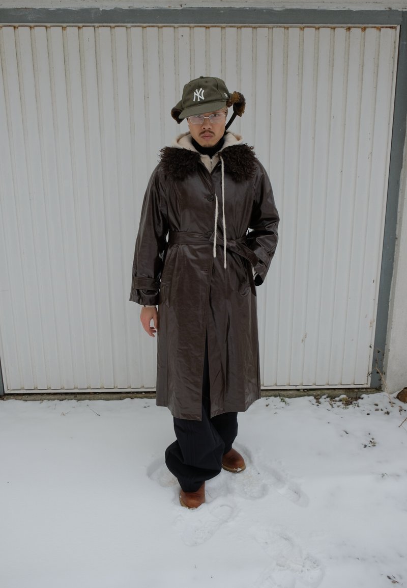 Person som bär en mörkbrun kappa med bälte och pälskrage, grön vintermössa med öronlappar, glasögon, står på snö framför en vit garageport.