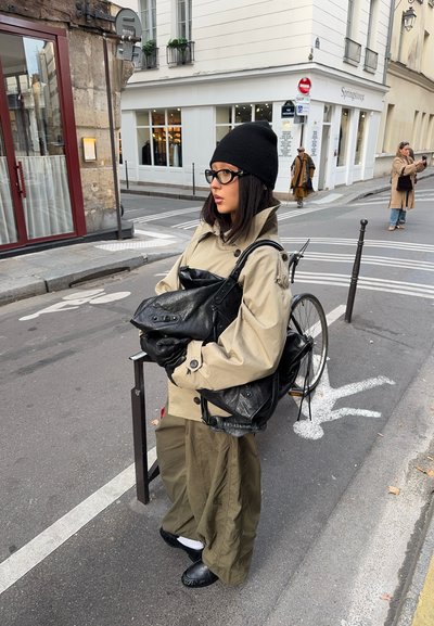 Mujer con gorro de lana y chaqueta beige sosteniendo un bolso de cuero negro junto a una bicicleta en una calle de la ciudad con paso de peatones y edificios.