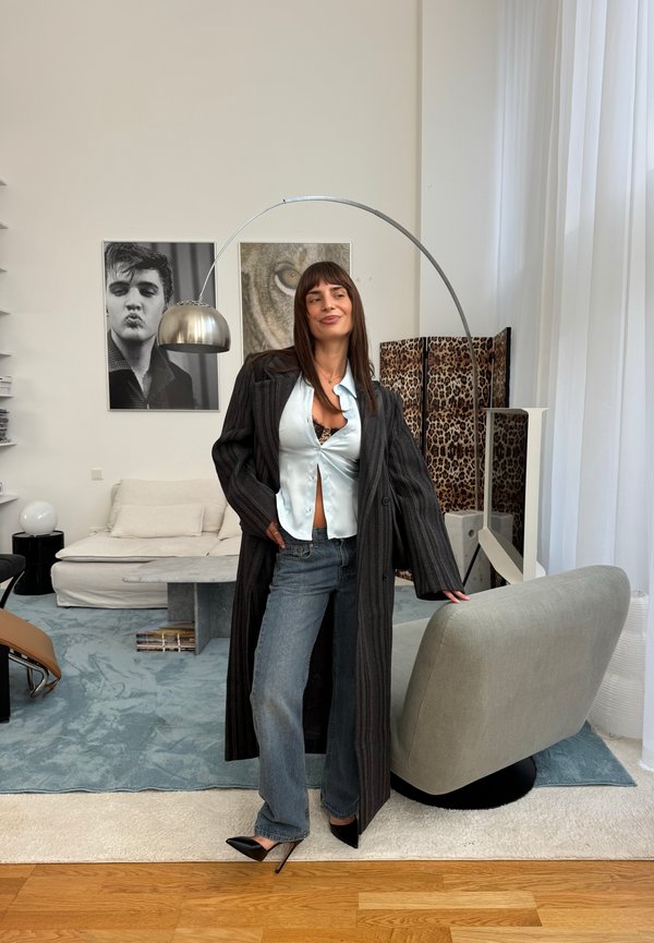 Une femme portant un long manteau sombre, un chemisier blanc, un jean et des talons pose à côté d’une chaise dans un salon moderne.