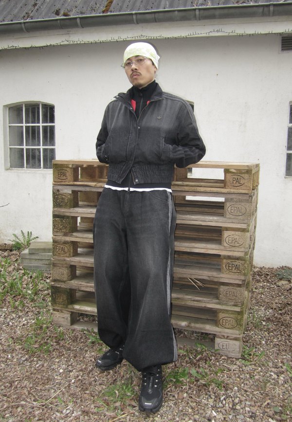 Personne portant une veste noire, un pantalon large noir et un foulard clair sur la tête, se tient les mains dans les poches devant des palettes en bois empilées.