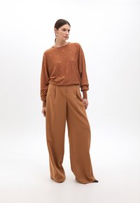Hnedý sveter so zlatými kruhovými akcentmi a voľným strihom, kombinovaný s vysokými, širokými nohavicami v beige farbe. Hladká textúra a uvoľnená silueta.