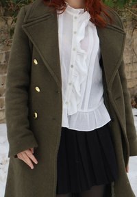 Femme portant un manteau vert foncé avec des boutons dorés sur un chemisier blanc à volants et une jupe plissée noire, debout sur un sol recouvert de neige.