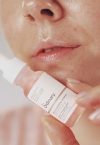 Nærbilde av en person som holder en flaske med The Ordinary hudpleieserum nær leppene, med glatt hud og en nesering.