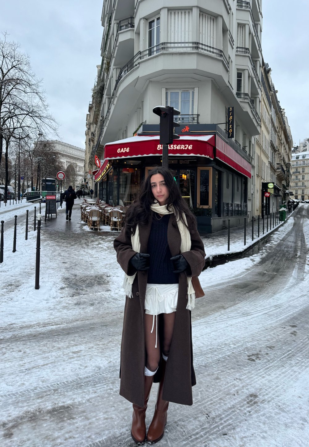 Jeune femme portant un long manteau, une écharpe et des bottes, debout à un coin de rue enneigé à Paris, près d'un café-brasserie aux auvents rouges.
