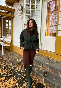 Donna con giacca verde, leggings marroni e stivali da pioggia verdi in piedi su scalini di pietra coperti di foglie davanti a una casa bianca e gialla.