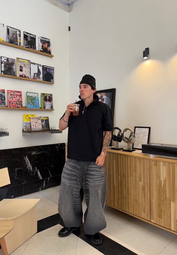 Joven con un gorro negro y pantalones grises anchos bebe café frío en una habitación moderna con revistas y auriculares en exhibición.