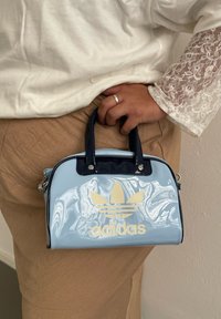 Απαλή μπλε πατενταρισμένη ψεύτικη δερμάτινη τσάντα με σκούρους μπλε λαβές και χρυσό λογότυπο adidas. Διαθέτει στρογγυλή μορφή με γυαλιστερή επιφάνεια.