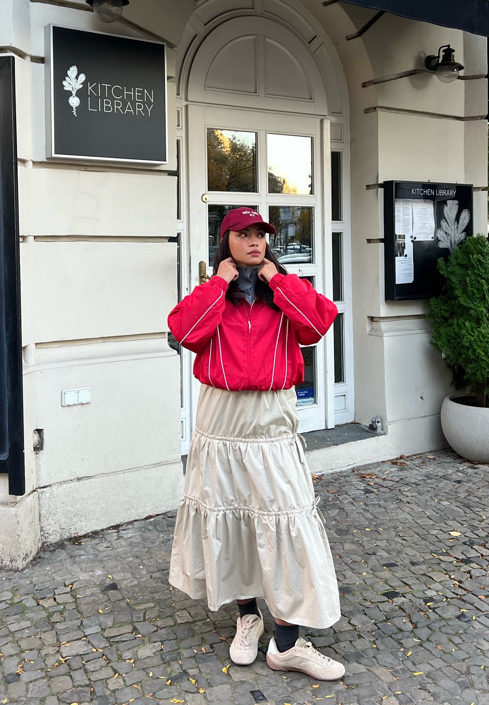 Rød bomberjakke med hvide kanter, cremet lagdelt nederdel og hvide sneakers. Tilbehør inkluderer en bordeaux kasket. Baggrunden viser en caféindgang.