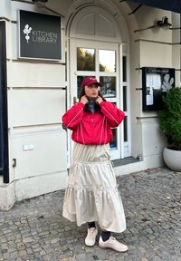 Rød bomberjakke med hvide kanter, cremet lagdelt nederdel og hvide sneakers. Tilbehør inkluderer en bordeaux kasket. Baggrunden viser en caféindgang.