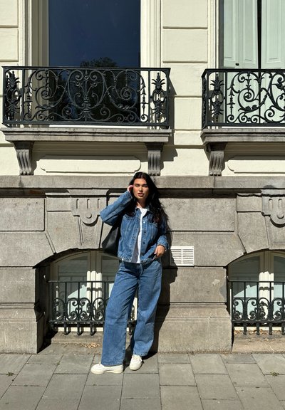 Conjunto de denim que incluye una chaqueta y pantalones de pierna ancha en azul claro. Bolsa tote negra, zapatos blancos, de pie frente a una pared de piedra decorativa.