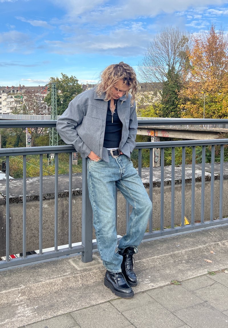 Grey cropped jacket over a black top, light blue baggy jeans, and black chunky boots. Person leans against a railing outdoors.