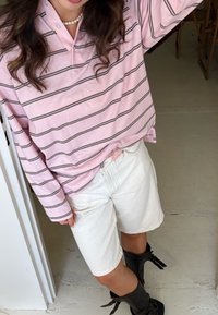 Camisa de manga comprida rosa clara com riscas cinzentas, combinada com calções brancos. Botas altas pretas com detalhe em fivela. Roupa casual.