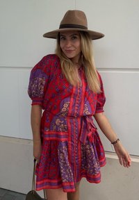 Rochie florală roșie și mov cu mâneci bufante, detalii cu butoni și talie legată. Însoțită de o pălărie maro cu boruri largi și o geantă.