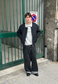 Giacca bomber nera sopra un maglione grigio chiaro, abbinata a jeans larghi scuri e scarpe nere. In piedi vicino a un cancello verde.