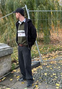 Czarna bluza z kapturem na zamek, założona na zieloną koszulę polo z białym paskiem, zestawiona z luźnymi czarnymi dżinsami oraz czarnymi trampkami z szarymi akcentami.