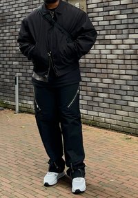 Black bomber jacket with a zip, dark shirt underneath, black trousers with zippered pockets, and white trainers with black accents.