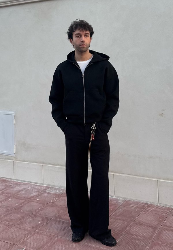 Črn zip-up hooded pulover z gladko teksturo, v kombinaciji z ohlapnimi črnimi hlačami. Opazna podrobnost: ključi na ključavnici pritrjeni na hlače.
