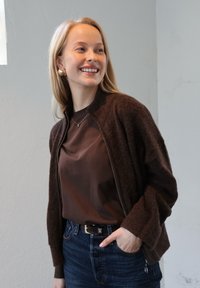 Hnedý kardigán na zips s jemnou textúrou, skombinovaný s hnedou košeľou a džínami. Pohľad dopĺňajú zlaté náušnice a jednoduchý náhrdelník.