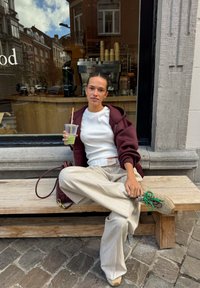 Persoon die op een houten bankje zit buiten een café, gekleed in een kastanjebruine hoodie, een witte top en een beige wijde broek, terwijl hij/zij een drankje vasthoudt.