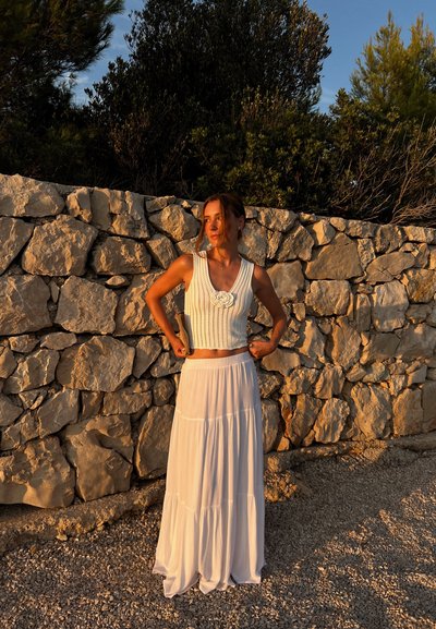 Top corto de punto blanco con detalles de flores, combinado con una falda maxi blanca de capa fluida. Fondo de pared de piedra natural, luz de la tarde.
