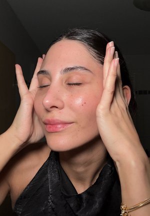 Gros plan sur un visage avec une peau lisse, de légères taches de rousseur et les yeux fermés. Les mains sont posées sur les joues. La personne porte un haut noir texturé.